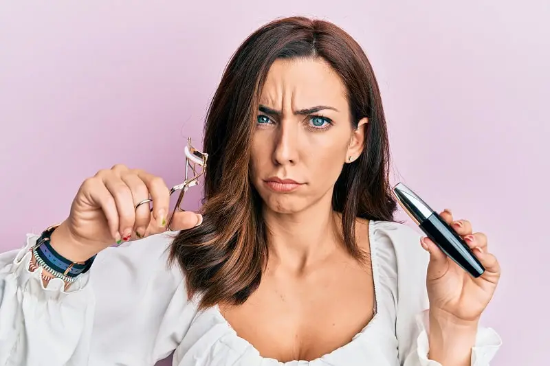 young woman holding an eyelash curler and mascara skeptical about curling her lashes without mascara